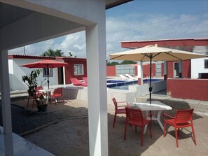 An outdoor patio with red chairs and a table, a pool area with lounge chairs, and a building with a red roof and white walls.