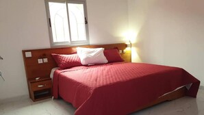 A bedroom with a bed, wooden headboard, bedside tables, a nightstand, a window, and a wall-mounted light.