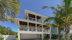 A two-story building with a covered balcony, a garage, and a well-maintained garden with palm trees. - Footprints in the Sand managed by Beach Retreats (Holmes Beach)