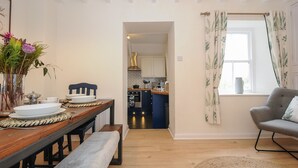 A modern kitchen with white cabinets, a blue countertop, and a visible stove. There is a dining area with a wooden table, benches, and a vase with flowers. A grey armchair and a window with curtains are also visible. - Carvannel Cottages (Camborne)