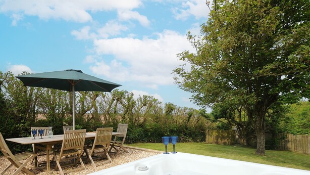 A hot tub with a table and chairs, an umbrella, and a grassy yard with trees. - Carvannel Cottages (Camborne)
