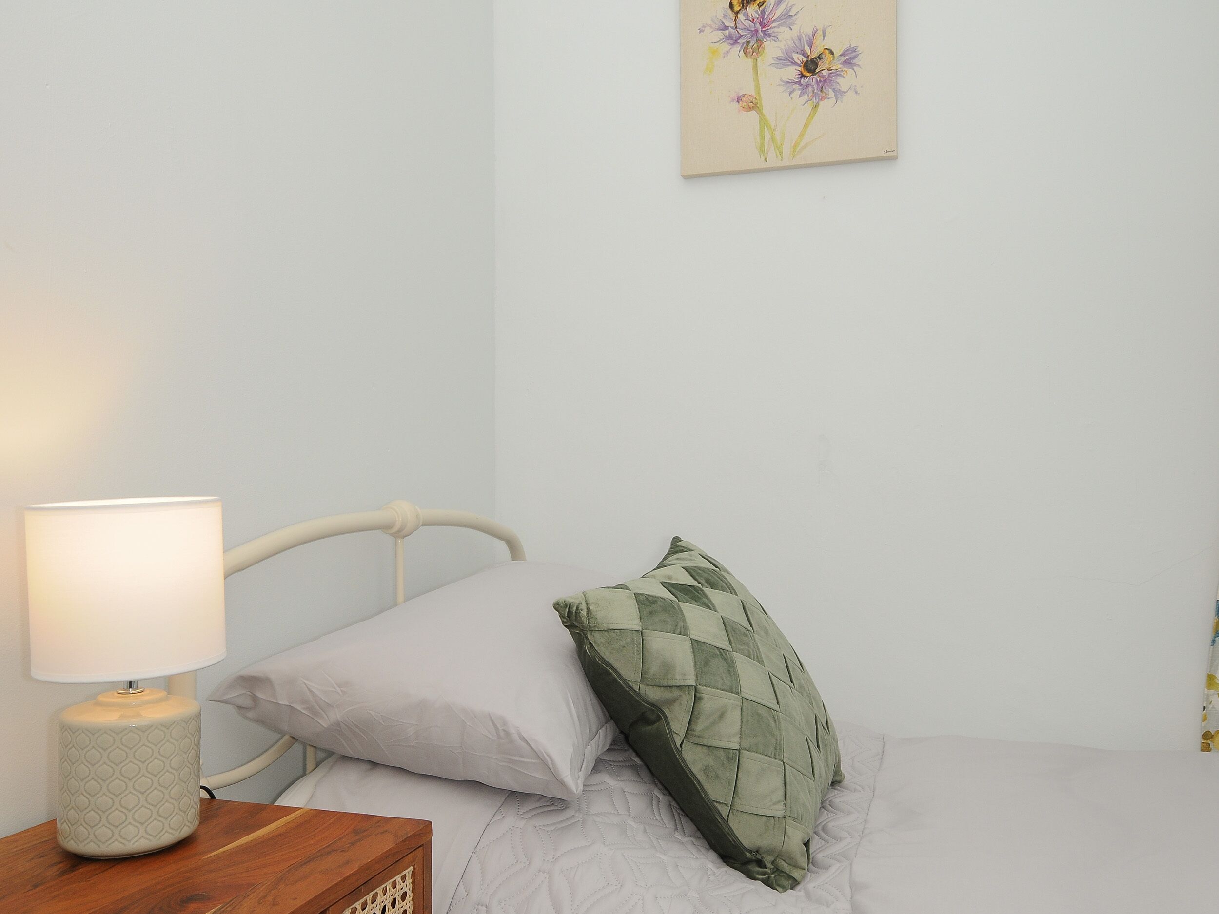 Un lit avec un oreiller blanc et un oreiller vert matelassé, une table de chevet avec une lampe, et un tableau représentant des fleurs accroché au mur.