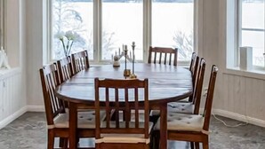 Een ronde eettafel met zes houten stoelen, grote ramen met uitzicht op een besneeuwd landschap, en een radiator aan de muur.