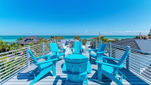 Una terraza de madera con sillas turquesas y una mesa redonda, con vista a una playa de aguas azules cristalinas y palmeras.