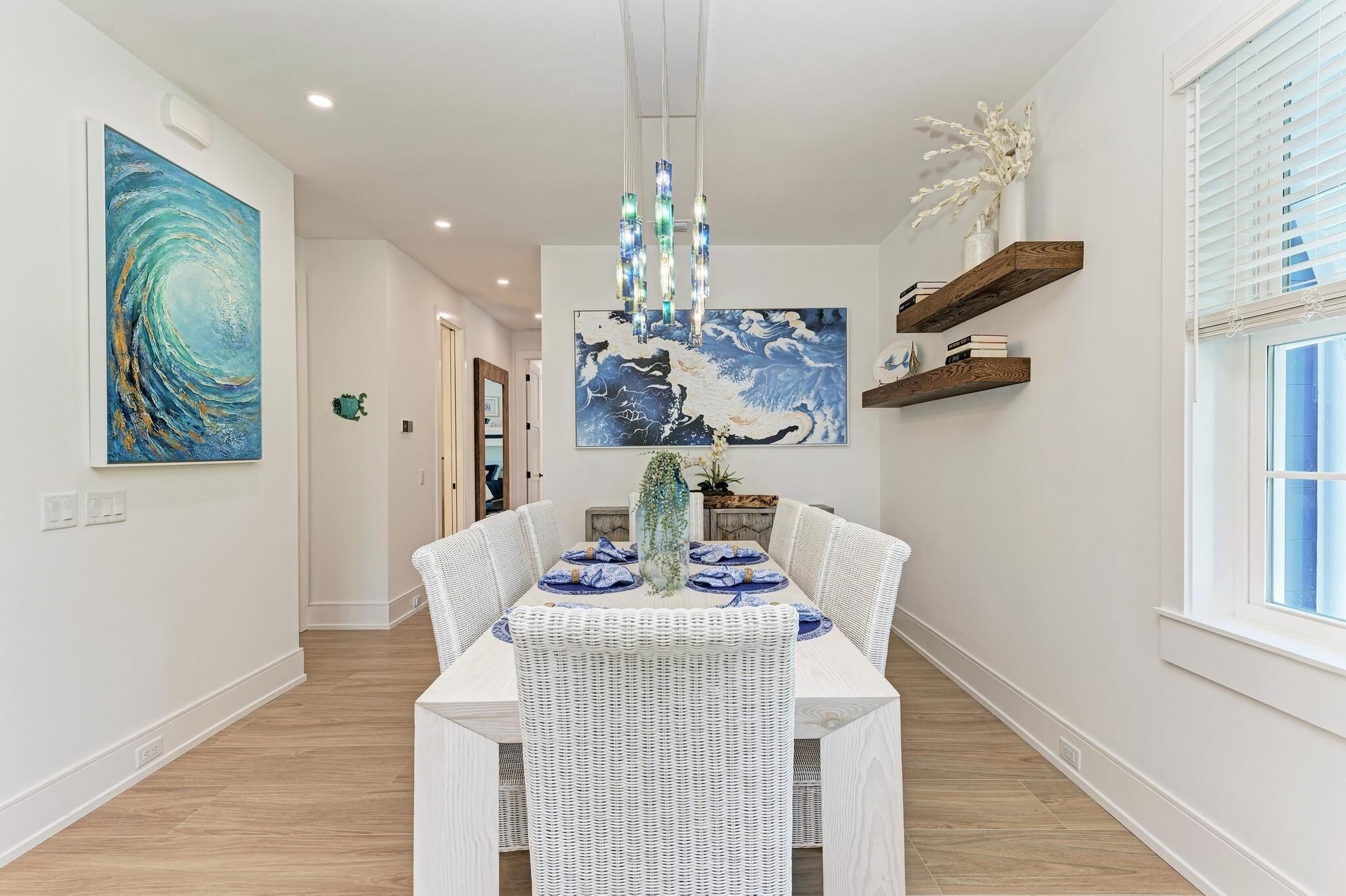 Un coin repas avec une table blanche dressée pour quatre personnes, entourée de chaises en osier, d’un luminaire suspendu et d’œuvres d’art accrochées aux murs.