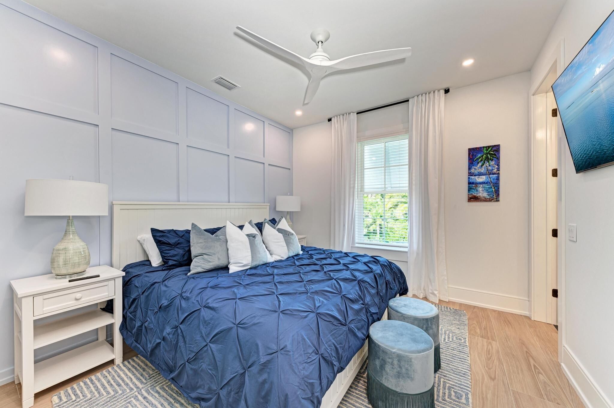A bedroom with a bed, nightstand, lamp, ceiling fan, window with curtains, and a painting on the wall.