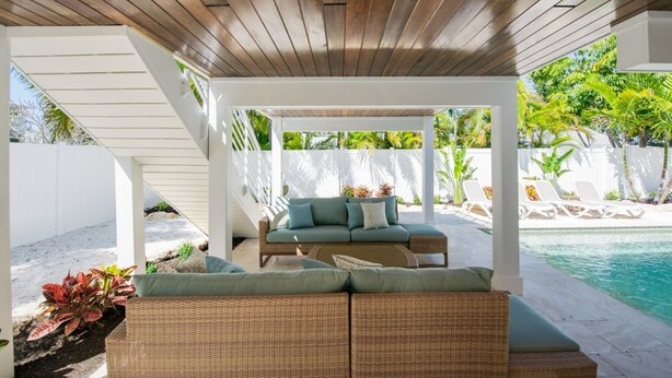 Ein überdachter Außenbereich mit Rattan-Sofa und Kissen, im Hintergrund ein Pool und ein weißer Zaun, der den Bereich umgibt.