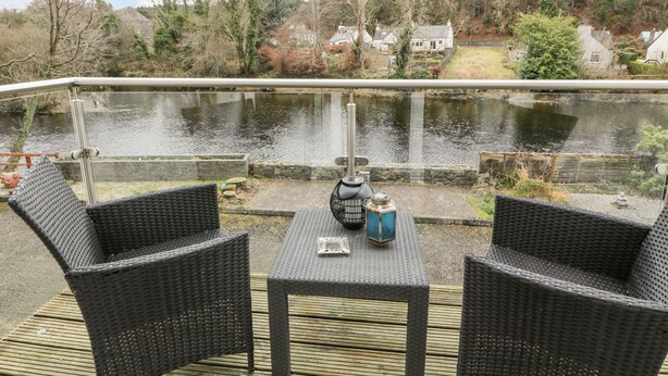 A balcony with wicker furniture and a view of a river and houses in the background. - Barley Cottage (Newton Stewart)