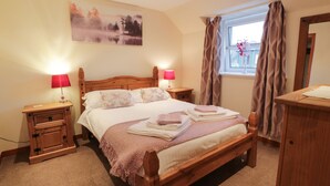 A neatly arranged bedroom with a wooden bed, two bedside tables with lamps, a window with curtains, and a painting on the wall. - Barley Cottage (Newton Stewart)