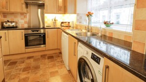 A modern kitchen with wooden cabinets, stainless steel appliances, a built-in oven, a dishwasher, and a window with a view of the outside. - Barley Cottage (Newton Stewart)