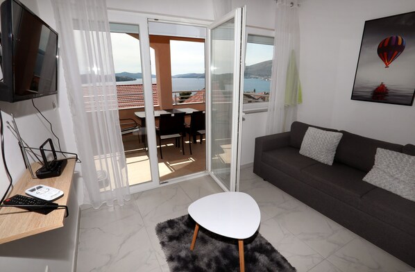 A modern living room with a sofa, a small white table, a TV, and a balcony with a view of the sea.
