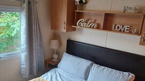 A bedroom with a bed, a nightstand, a lamp, and a window with curtains. - Daisy's Luxury Caravan (Lincoln)