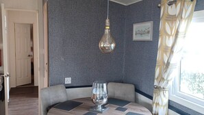 A dining area with a round table, chairs, a hanging light fixture, and a window with curtains. - Daisy's Luxury Caravan (Lincoln)
