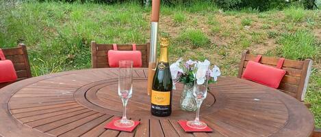 A round wooden table with a red umbrella, surrounded by wooden chairs with red cushions. A bottle of champagne and two glasses are placed on the table.