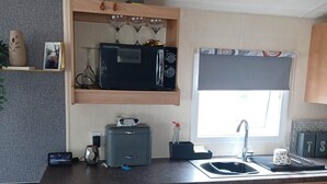 A compact kitchen with a microwave, toaster, and sink. There is a small window with a curtain, a shelf with decorative items, and a plant. - Daisy's Luxury Caravan (Lincoln)