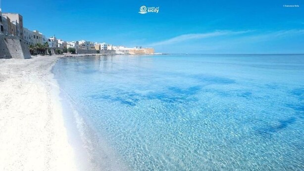 Una playa con agua azul cristalina y arena blanca, edificios a lo largo de la costa, y un cielo despejado.