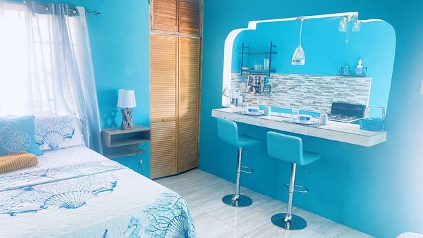 A bedroom with a bed, a desk, two chairs, a wardrobe, and a bathroom with a mirror and sink. - YanceyLargo Estate - Newly Built Modern 1 Bedroom Suite 1 Villa by RedAwning (Montego Bay)