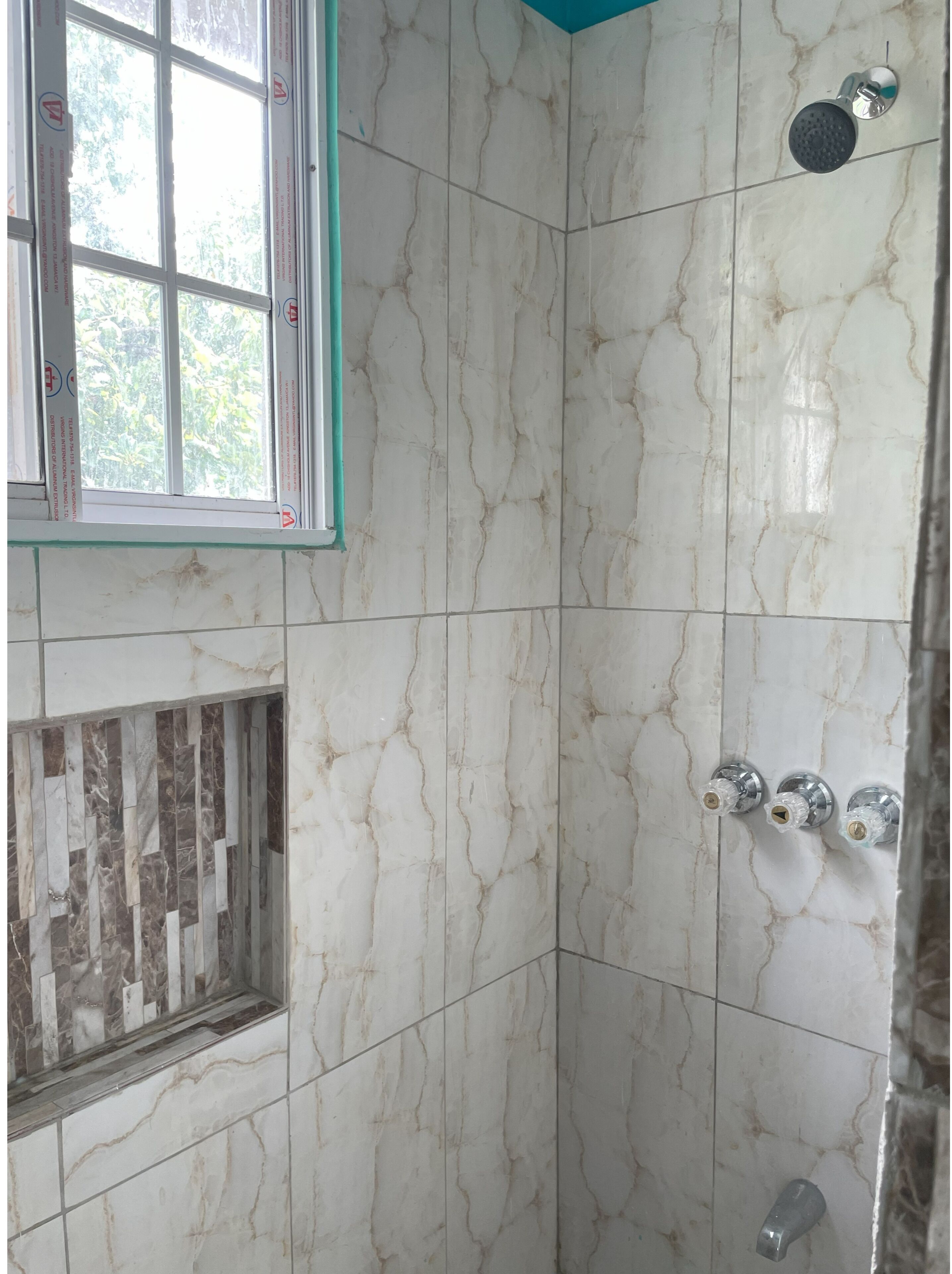 Un baño con baldosas de mármol blanco, un cabezal de ducha y una ventana con vistas a la vegetación.