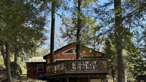 Een rustieke chalet met een houten terras, omgeven door bomen en een stenen pad dat naar de ingang leidt.
