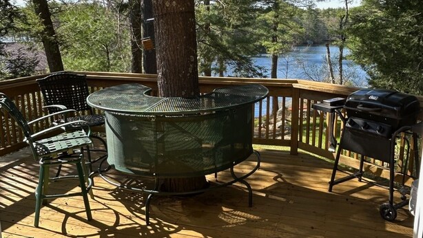 Een houten terras met een metalen barbecue en een metalen tafel met stoelen, met uitzicht op een meer.