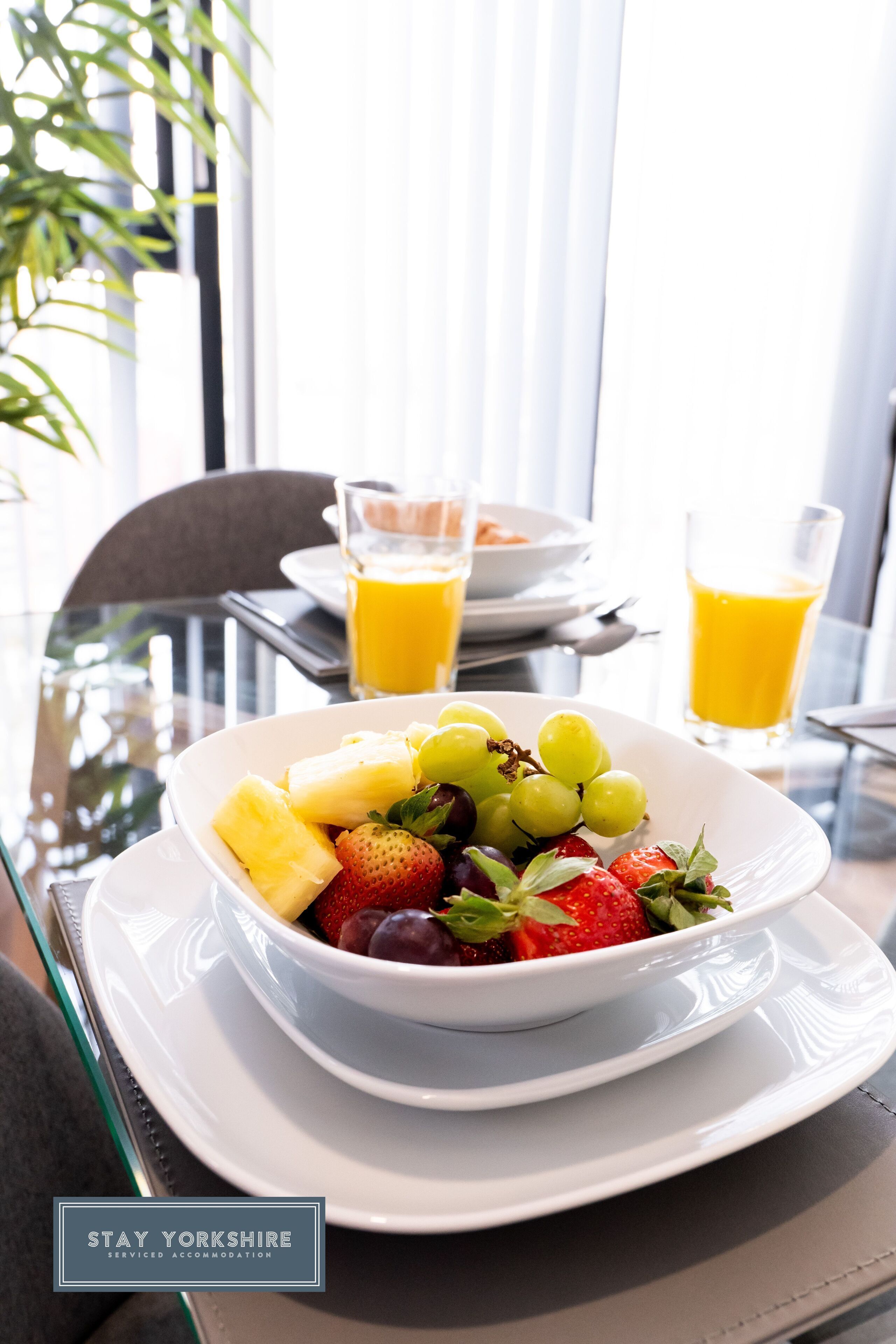 Een ontbijtsetting met een fruitschaal, twee glaasjes sinaasappelsap en een bord met toast op een tafel.