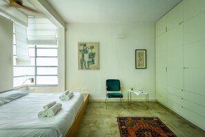 Quarto de hotel com uma cama, uma cadeira, uma mesinha, um quadro e um tapete estampado.