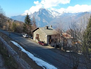 Un bâtiment en pierre avec un toit en pente, une voiture garée devant et une route menant à une chaîne de montagnes.