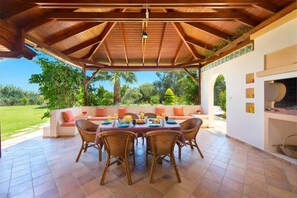 A covered outdoor dining area with wicker chairs and a table set for breakfast, overlooking a lush garden and palm trees. - Stunning 5-bed Villa in Gennadi! (Rhodes)