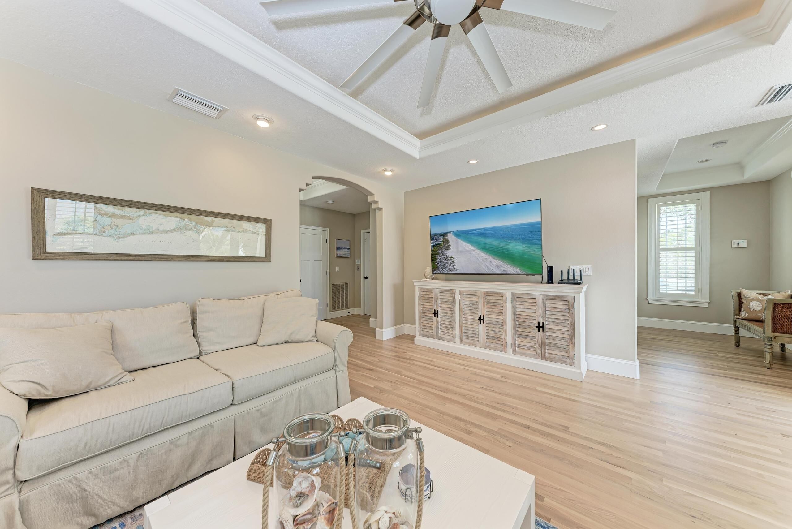 A spacious living room with a light-colored sofa, a coffee table with decorative jars, a television on a white cabinet, and a large painting on the wall.