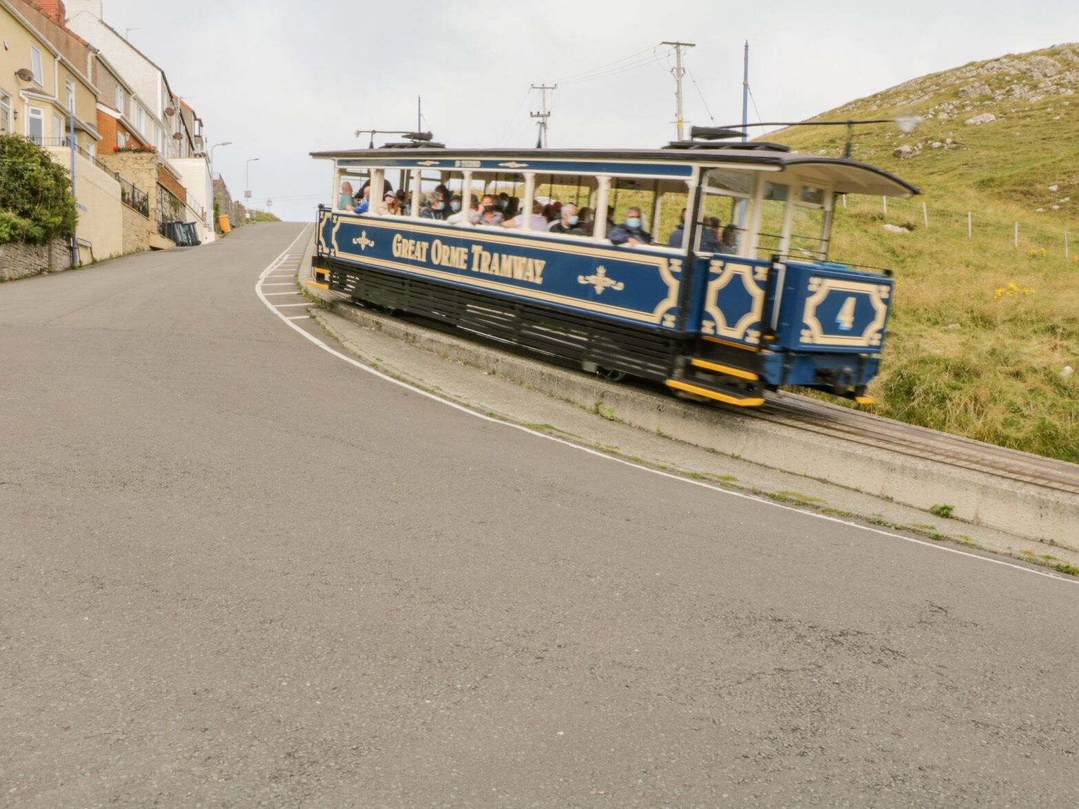 A Great Orme Tramway with passengers, traveling on a road with buildings on one side.