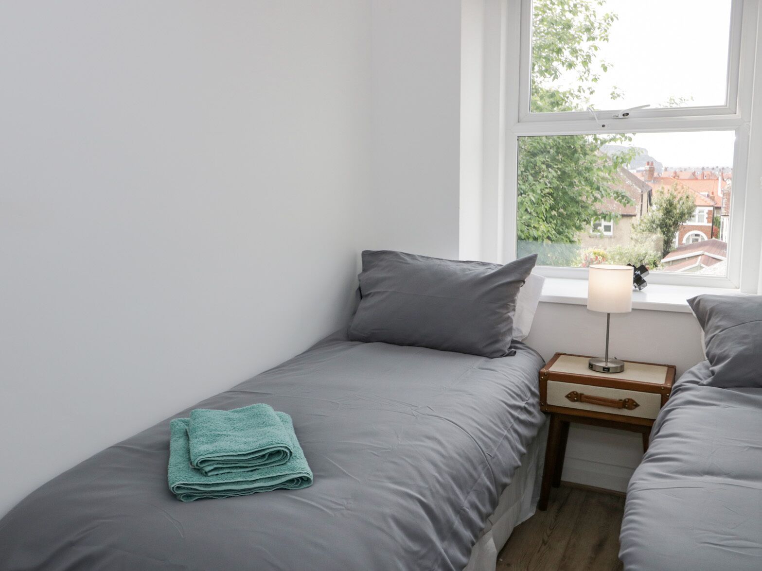 Une petite chambre propre et bien rangée, avec deux lits, une fenêtre donnant sur des immeubles et une table de chevet avec une lampe.