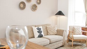 A living room with a sofa, a wooden side table, a glass of wine, and decorative wall art. - Ty Arth (Llandudno)