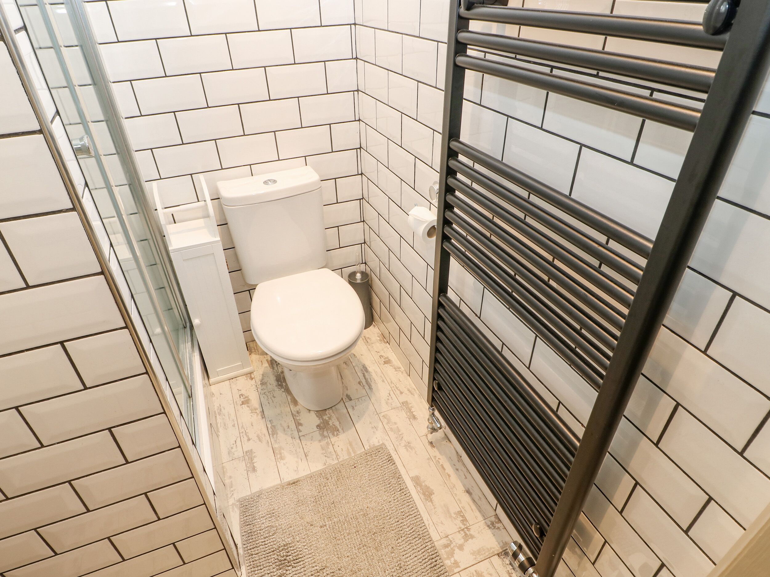 A small, modern bathroom with white tiles, a toilet, a glass shower enclosure, and a towel rack.