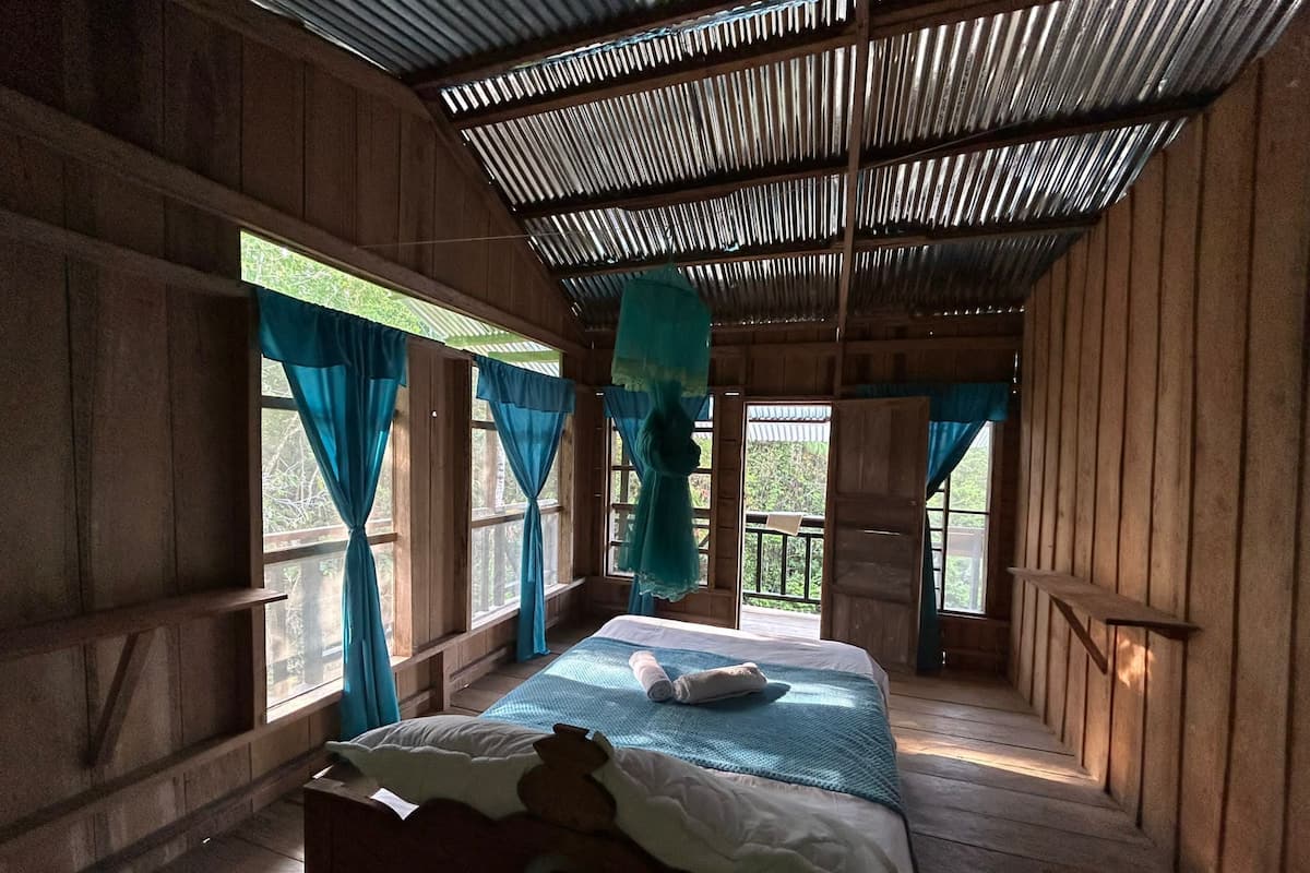 A wooden room with a bed, curtains, and a view of greenery.