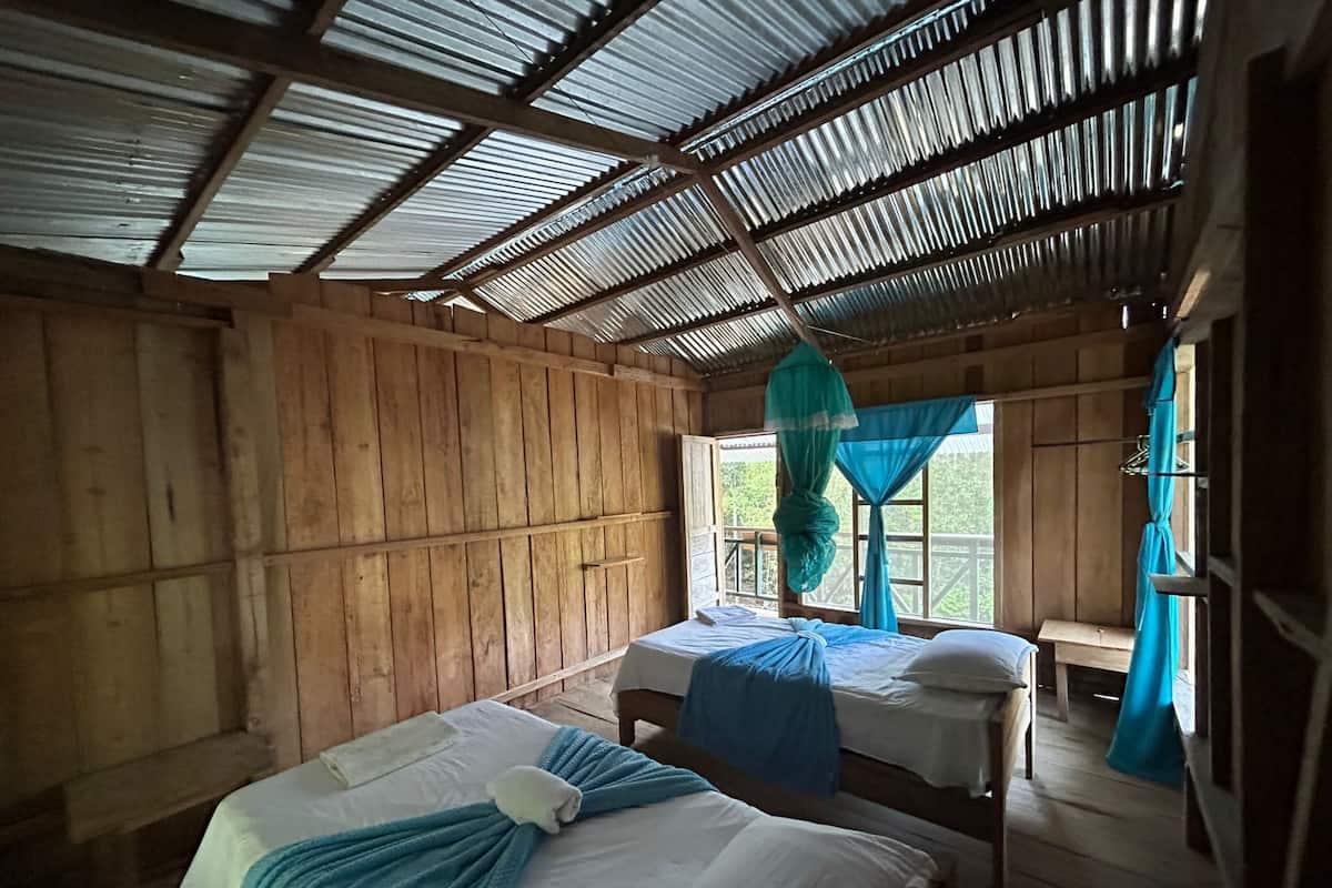 A rustic wooden room with two beds, a window with curtains, and a metal roof.