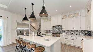 A modern kitchen with white cabinetry, a large island with bar stools, pendant lighting, and a stainless steel refrigerator. - Just Beachy managed by Beach Retreats (Holmes Beach)