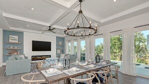 A spacious dining area with a wooden table set for six, surrounded by wooden chairs with cushioned seats. A large chandelier hangs from the ceiling, and a flat-screen TV is mounted on the wall. - Just Beachy managed by Beach Retreats (Holmes Beach)