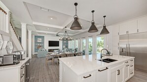 A modern kitchen with white cabinetry, a large island with a sink, pendant lighting, and a stainless steel refrigerator. - Just Beachy managed by Beach Retreats (Holmes Beach)