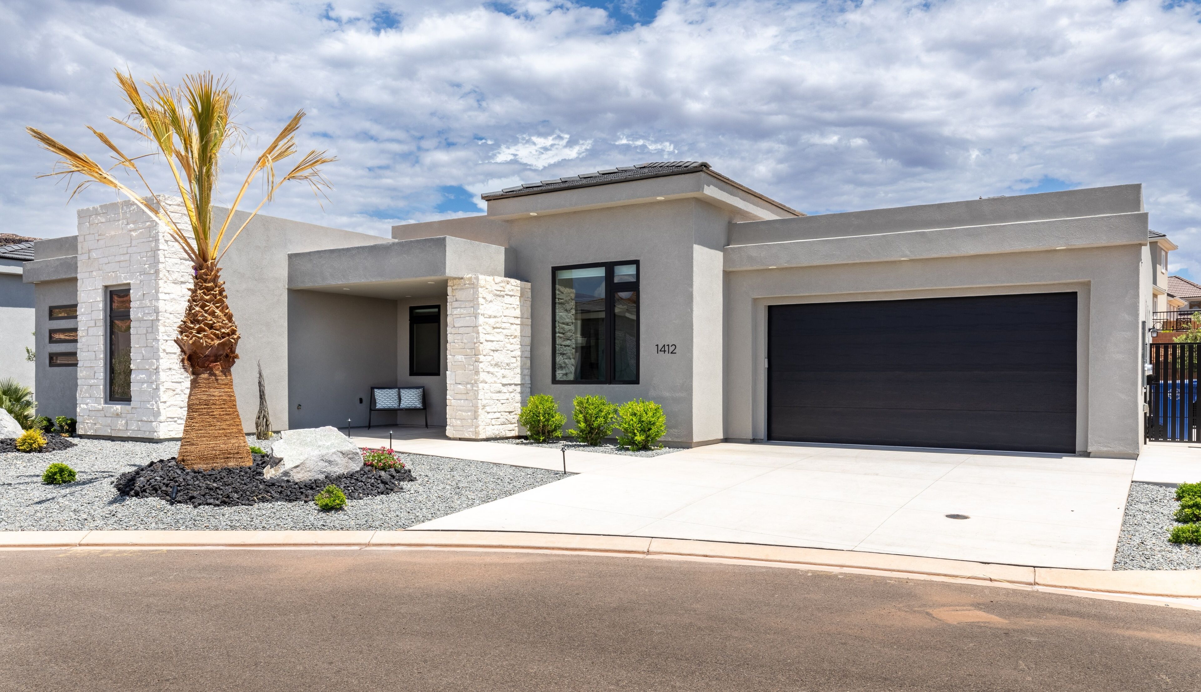 A modern two-story house with a garage, stone facade, and a palm tree in the front yard.