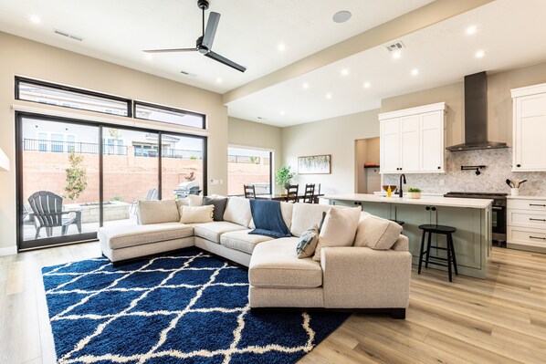 A modern living room with a sectional sofa, a blue patterned rug, and a kitchen area with white cabinets and a bar stool. - Sunshine Retreat 2 Bedroom Home by RedAwning (Washington)