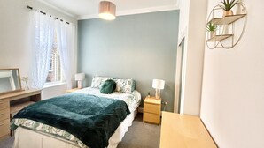 A bedroom with a bed, bedside tables, a dresser, a mirror, a window with curtains, and a wall-mounted shelf with plants. - Ground Floor Victorian Apartment (Coatbridge)