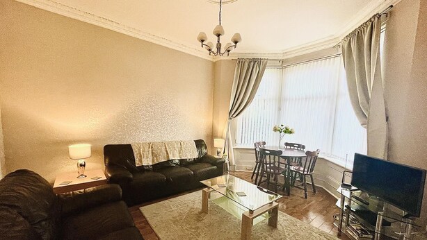 A living room with a sofa, coffee table, TV, and a dining area with chairs and a table. - Ground Floor Victorian Apartment (Coatbridge)