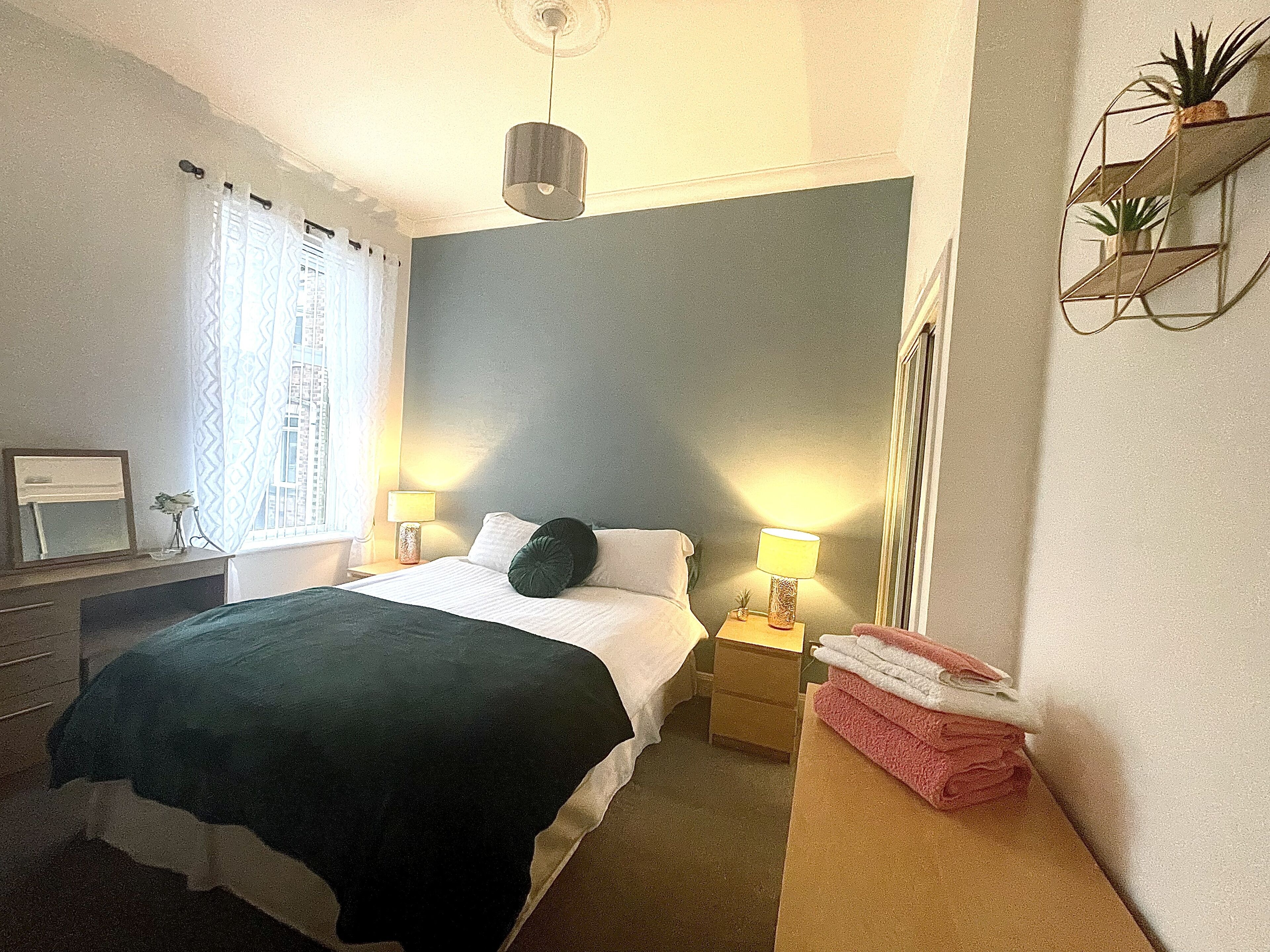 Ein ordentlich eingerichtetes Schlafzimmer mit Bett, Nachttischlampen, einem Nachttisch, einem Spiegel und einem kleinen Regal mit Pflanzen.