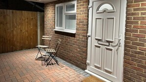 A brick exterior with a white door and window, a wooden fence, and a small outdoor seating area with a metal chair and table. - Luxury Detached Studio Apartment in Luton (Luton)