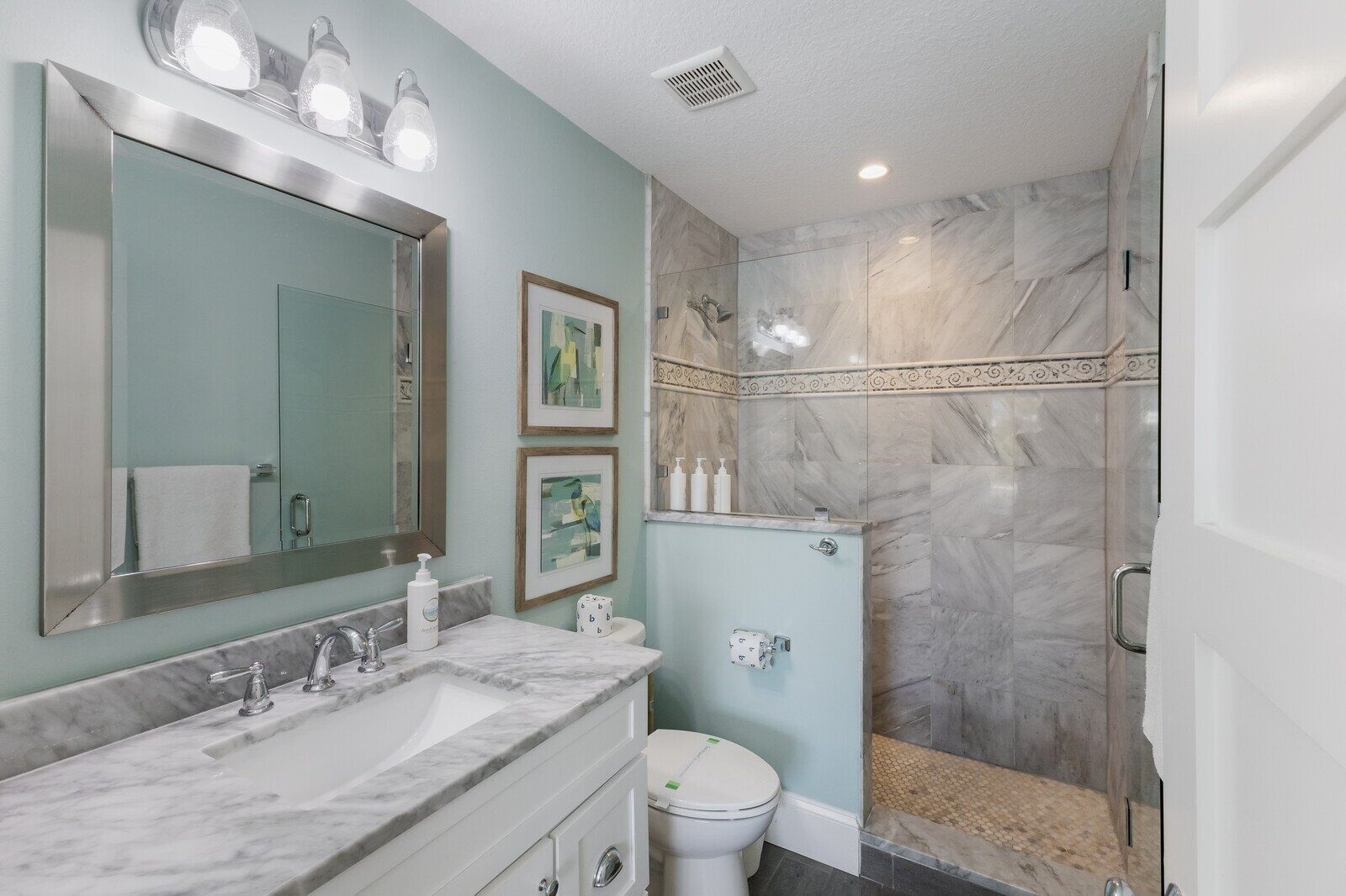 A bathroom with a marble sink, a large mirror, framed artwork, and a shower with marble tiles.