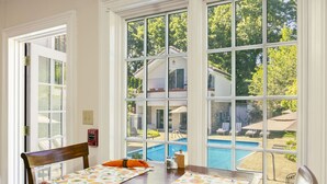 Un comedor con una mesa dispuesta para dos personas, ventanas amplias con vista a una piscina y una casa, y un detector de incendios en la pared.