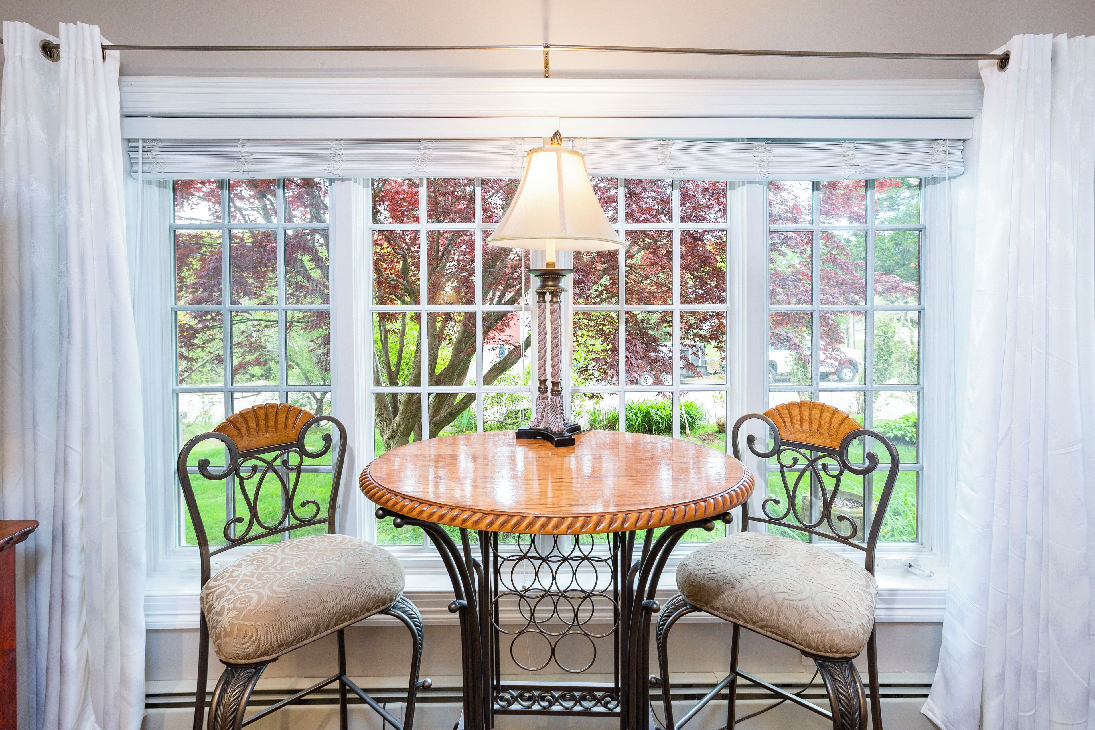 A dining area with a round table and two chairs, a lamp, and a view of trees through large windows.