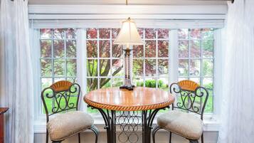 A dining area with a round table and two chairs, a lamp, and a view of trees through large windows.