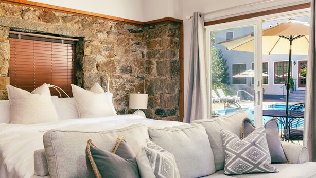 Un dormitorio con pared de piedra, una cama con muchos almohadones, una mesita de noche con lámpara y vistas a un área de piscina al aire libre.
