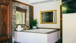 Un baño con una bañera grande, un mueble de madera, pared de piedra, planta en maceta y un espejo con marco dorado.
